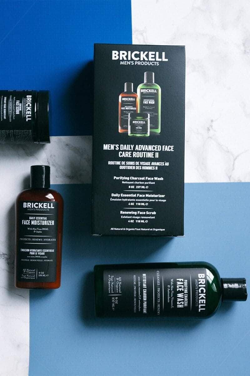 Brickell men's skincare products arranged on a blue and white surface, including a box for advanced face care routine, a bottle of face wash, a bottle of face scrub, and a bottle of face serum.
